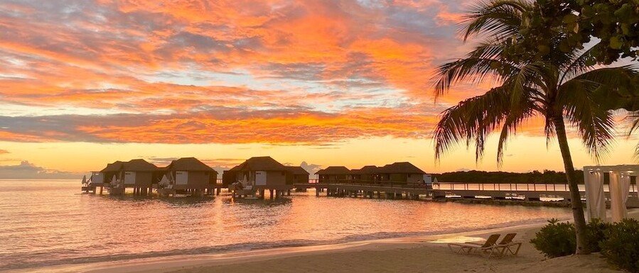 Sunset view of overwater villas at a Sandals resort with palm tree, beach chairs, and calm Caribbean water