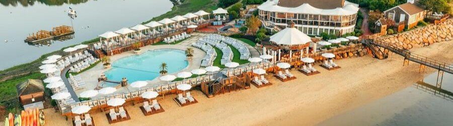 A resort pool area on a sandy beach with rows of lounge chairs, umbrellas, and a circular pool by the water's edge.