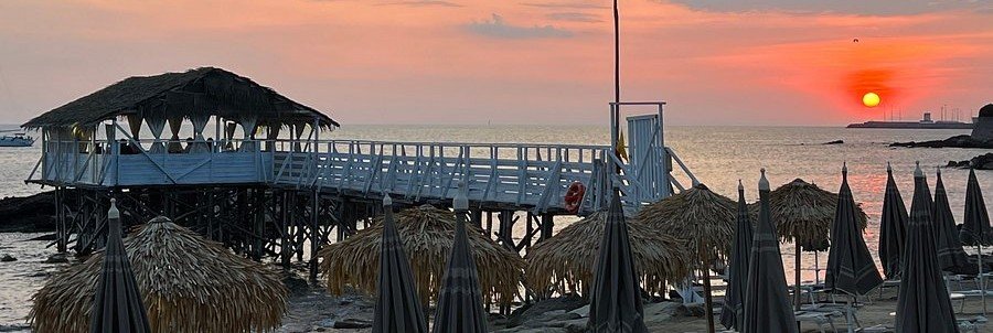 A coastal sunset view of the Grotta Aurelia beach club in Civitavecchia, featuring a thatched-roof overwater dining pavilion, a wooden pier, and closed straw beach umbrellas along the shore.