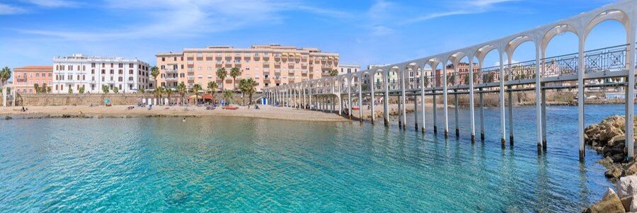 A wide panorama of the historic Il Pirgo pedestrian pier stretching across turquoise waters toward the central pebble beach and pastel-colored waterfront buildings of Civitavecchia.