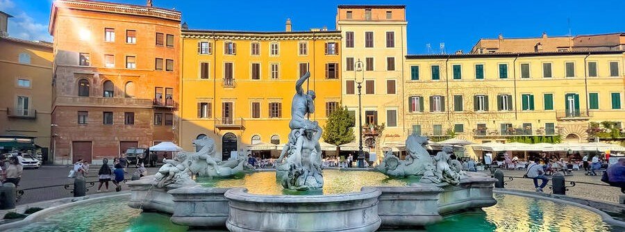 A sunlit panoramic view of the Fountain of Neptune in Rome's Piazza Navona, featuring detailed marble sculptures, spraying water, and historic pastel-colored buildings in the background.
