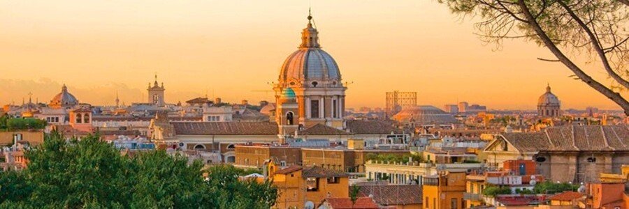 A wide panorama of the Rome cityscape at sunset, highlighting the grand Baroque dome of San Carlo al Corso and a golden horizon blanketed by historic tiled rooftops.