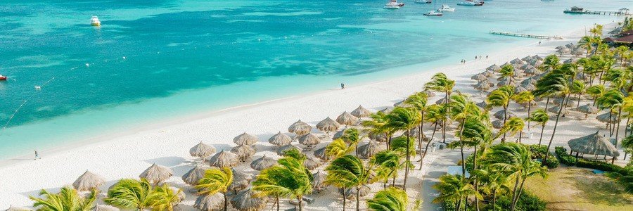 Aerial view of Palm Beach in Aruba showing turquoise Caribbean waters, white sand beach, palm trees, and thatched palapas.