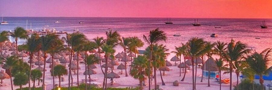 Vibrant pink and orange sunset sky over the ocean with palm trees and beach palapas at the Aruba Marriott Resort & Stellaris Casino.