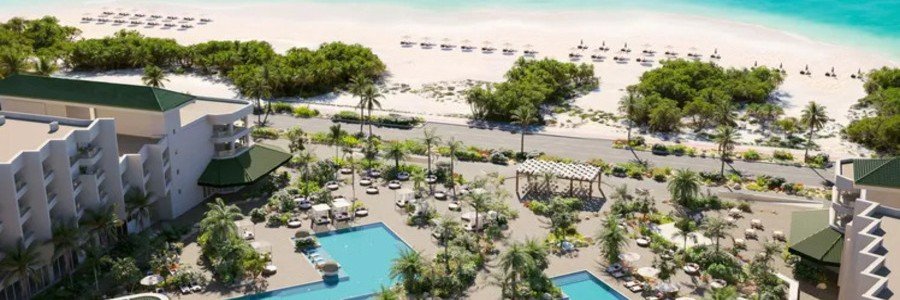 Aerial view of the low-rise buildings, outdoor swimming pool, lush courtyard gardens, and wide white sands of Eagle Beach at Manchebo Beach Resort & Spa.