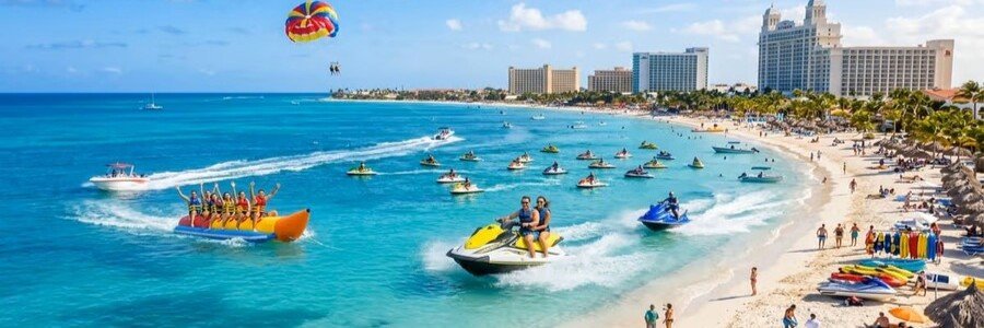 Tourists enjoying water sports at Palm Beach Aruba including jet skiing, a banana boat ride, and parasailing near high-rise resorts.