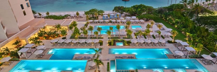 Aerial evening view of the luxury multi-tier outdoor swimming pools at The St. Regis Aruba Resort illuminated in blue next to the beach.