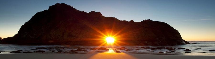 The glowing sun shines through the sea arch at Pfeiffer Beach in Big Sur, lighting the rugged cliffs with golden light.