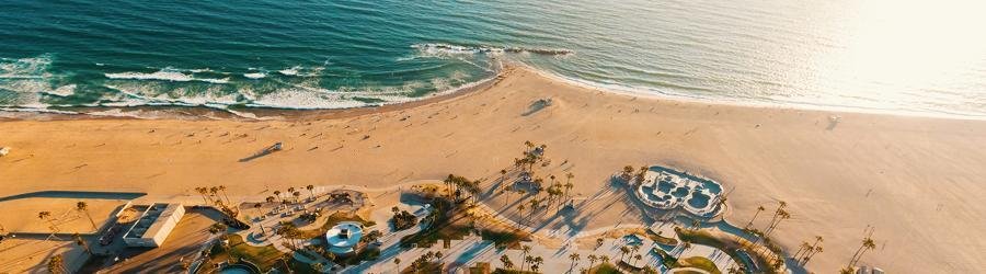 Aerial view of Venice Beach at sunset with palm trees, golden sand, and the iconic skate park beside the Pacific Ocean.