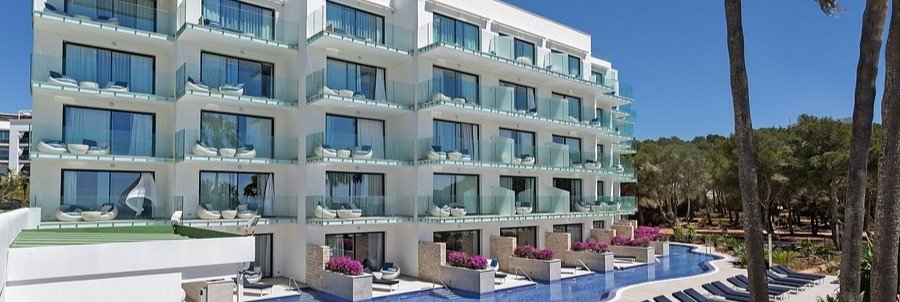 Exterior view of the contemporary white hotel block at Catalonia Royal Ses Estaques, showcasing sea-facing balconies and exclusive lower-level swim-up pool channels.
