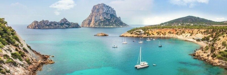 Turquoise coastal cove with sailboats and views of Es Vedrà near Ibiza surrounded by rocky cliffs and clear Mediterranean water.
