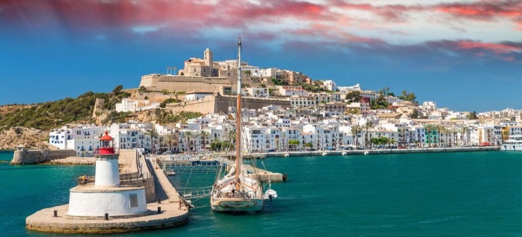 Dalt Vila overlooking Ibiza Town harbor with turquoise water, sailboats, lighthouse, and dramatic sunset clouds above the historic coastline.