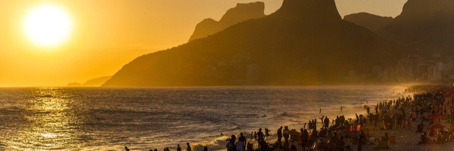 A crowded beach at sunset with a brilliant orange sun dipping over ocean waves next to massive mountain silhouettes.