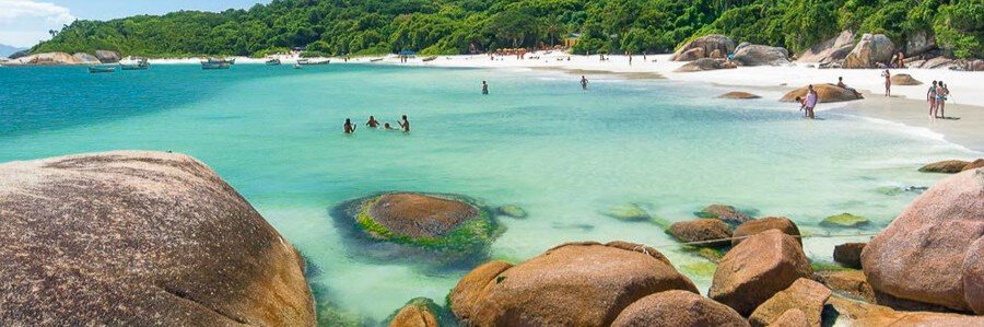 Large smooth boulders framing a crystal-clear turquoise bay with a white sand beach and lush green hills.