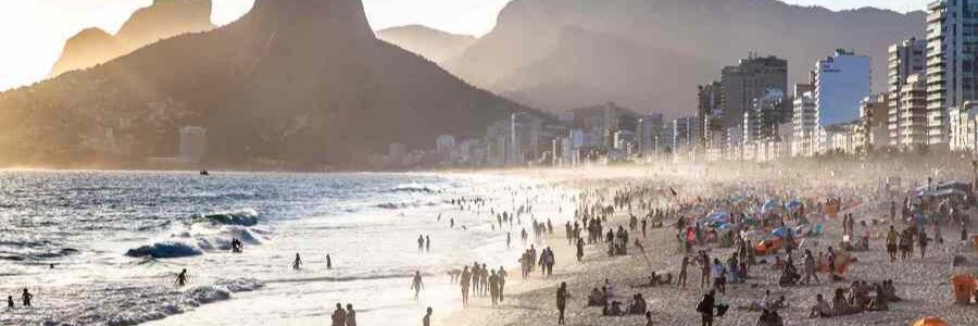 A crowded beach at sunset with ocean waves rolling onto the sand next to city buildings and massive mountain peaks.