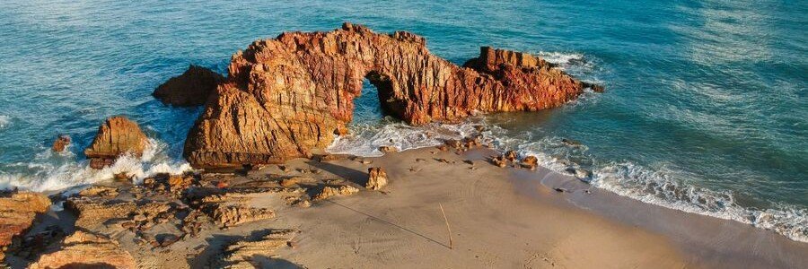 A natural rock arch formation standing on a golden sand beach as turquoise ocean waves crash around it.