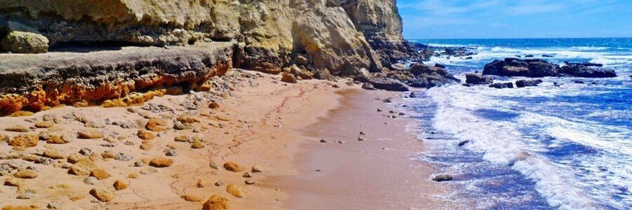 A rugged sandy beach lined with steep layered cliffs and deep blue ocean waves rolling onto the sand.