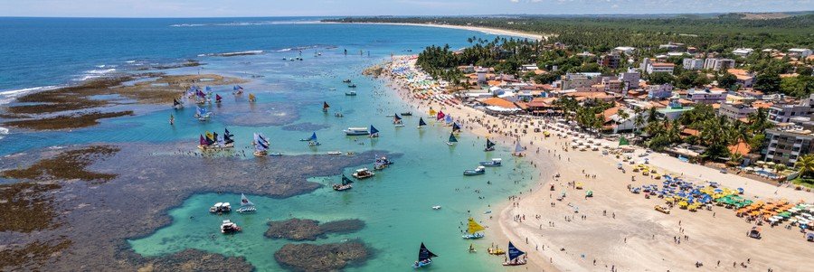 An aerial view of a wide sandy beach village next to flat coral reefs and turquoise natural tide pools.