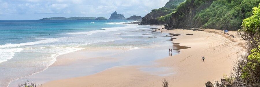 An expansive beach on a remote tropical island with deep blue ocean water, golden sand, and lush green cliffs.