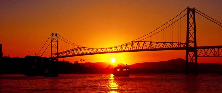 Hercílio Luz Bridge at Sunset Floripa | Beach Travel Destinations Hercílio Luz Bridge silhouetted against a colorful sunset over Florianópolis Bay.