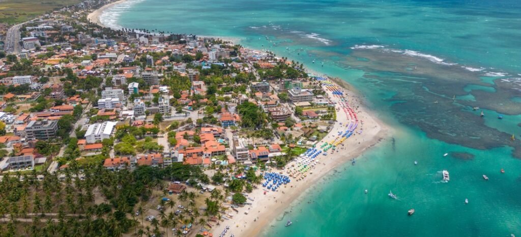 High-angle aerial view of Porto de Galinhas beach town in Brazil, showing rows of colorful beach umbrellas along the shoreline and coral reefs in the clear turquoise ocean.