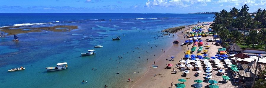 Aerial wide view of Porto de Galinhas beach showing traditional boats, coral reefs, and colorful umbrellas lining the shore.