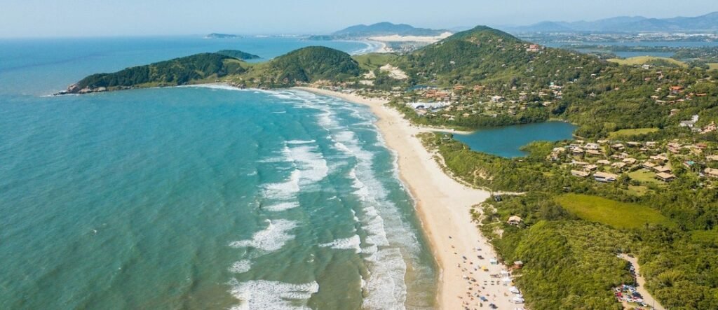 Aerial view of Praia do Rosa featuring a crescent-shaped beach, rolling waves, green hills, and coastal lagoons in Brazil