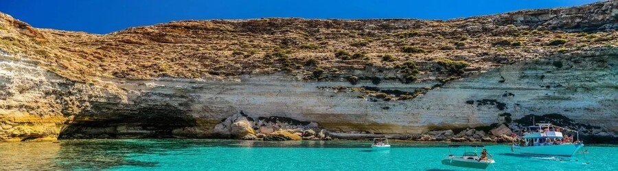 Turquoise water cove in Lampedusa with boats, swimmers, and limestone cliffs