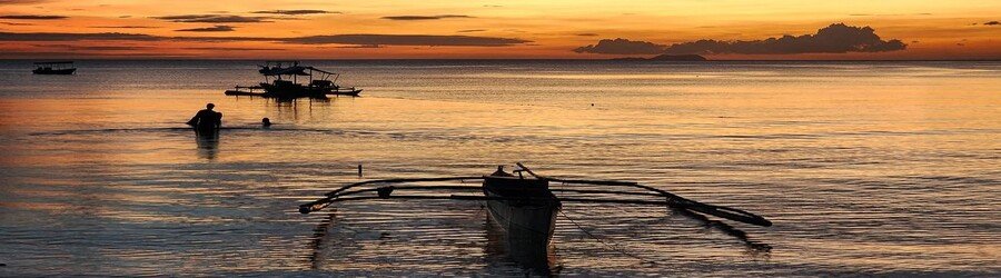 Sunset over the sea in Siquijor, Philippines, with calm water, traditional boats, and warm orange skies