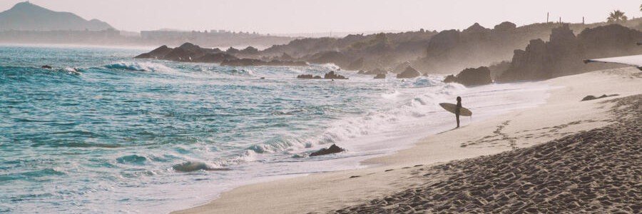 Cabo Beaches at Monuments Beach with a surfer walking beside rolling waves and dark rocks