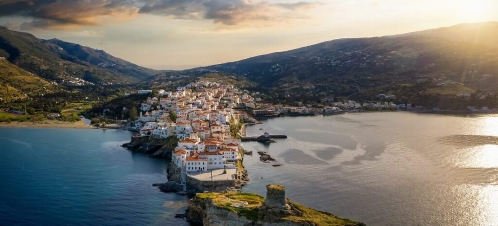 Aerial view of Chora, Andros, Greece, with whitewashed buildings stretching into the sea between two sheltered bays at sunset.