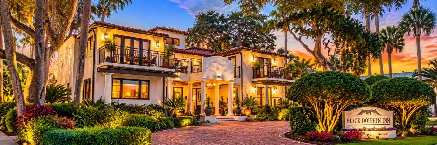 Black Dolphin Inn in New Smyrna Beach featuring Mediterranean-style architecture, illuminated balconies, lush tropical gardens, and a colorful sunset.