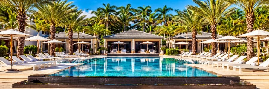 Luxury resort swimming pool surrounded by tropical palm trees, private cabanas, white lounge chairs, and shaded umbrellas.