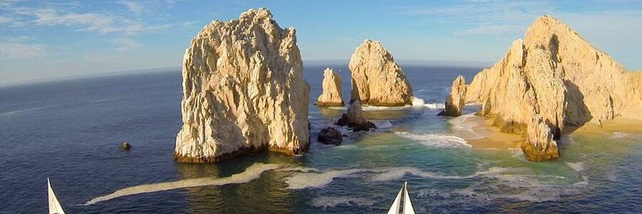 View of El Arco in Cabo San Lucas with large golden rock formations rising from the ocean, waves breaking around the base, and a sailboat in the foreground near Land’s End
