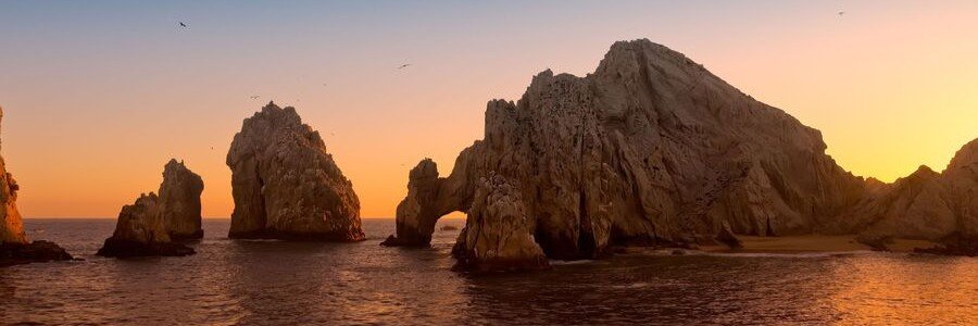 Sunset view of El Arco in Cabo San Lucas with glowing golden rock formations, calm ocean water reflecting warm colors, and birds flying above the Arch at Land’s End