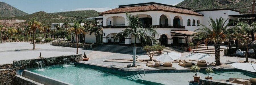 A luxury adults-only boutique hotel in La Paz featuring a large outdoor pool with lounge seating, palm trees, and a Spanish-style villa with arches and balconies set against a desert mountain backdrop.