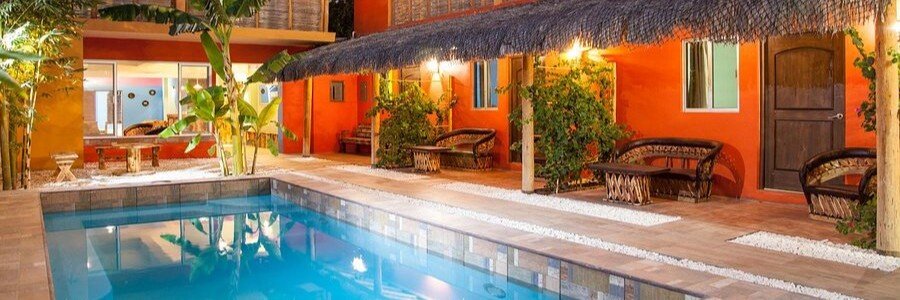 Cozy courtyard at Casa Juarez Bed and Breakfast in La Paz featuring a small outdoor pool, colorful orange walls, thatched roof details, and tropical plants with seating areas outside each room.