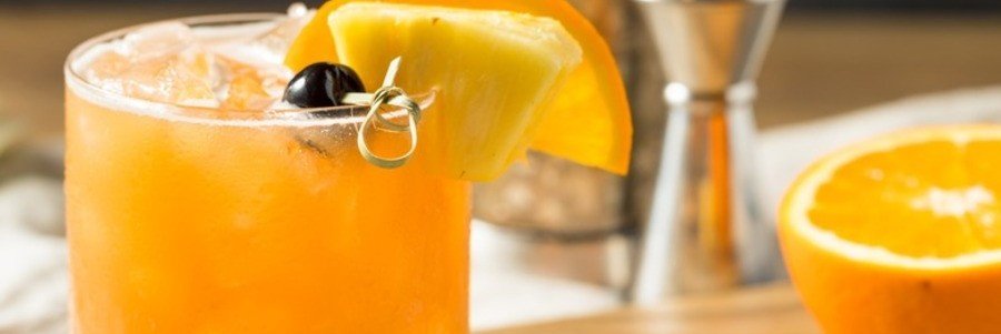 A close-up shot of a bright orange Rum Runner cocktail over crushed ice, garnished with a pineapple wedge, orange slice, and a cherry.