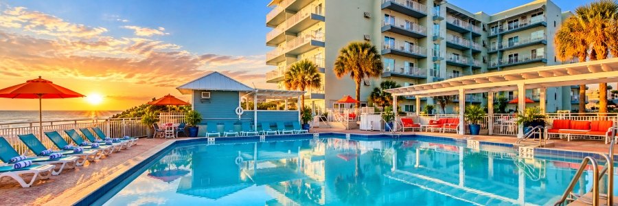 Oceanfront swimming pool at Coconut Palms Beach Resort II, featuring turquoise water, colorful umbrellas, palm trees, and sunset views.