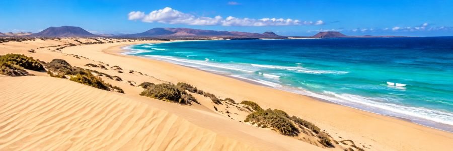 Panoramic view of Grandes Playas de Corralejo, featuring golden sand dunes, turquoise Atlantic waters, and volcanic mountains in Fuerteventura.