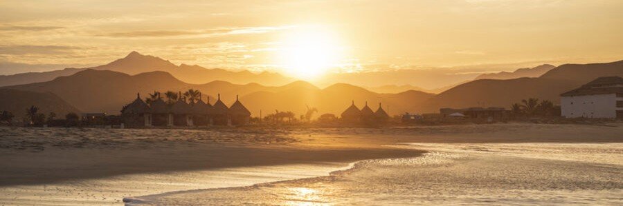 Sunset over Todos Santos beach with golden sky, ocean waves, distant mountains, and coastal buildings in Baja California Sur Mexico