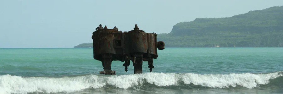 Weathered offshore structure surrounded by waves along a green and mountainous stretch of Haiti’s coastline.