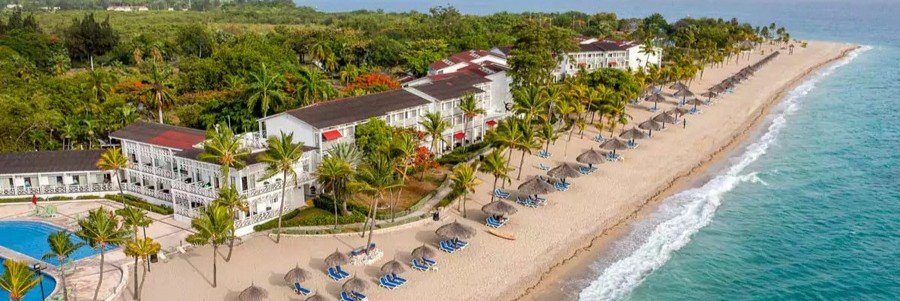 Aerial view of a palm-lined beach resort with pools, lounge chairs, sandy shoreline, and turquoise Caribbean water in Haiti.