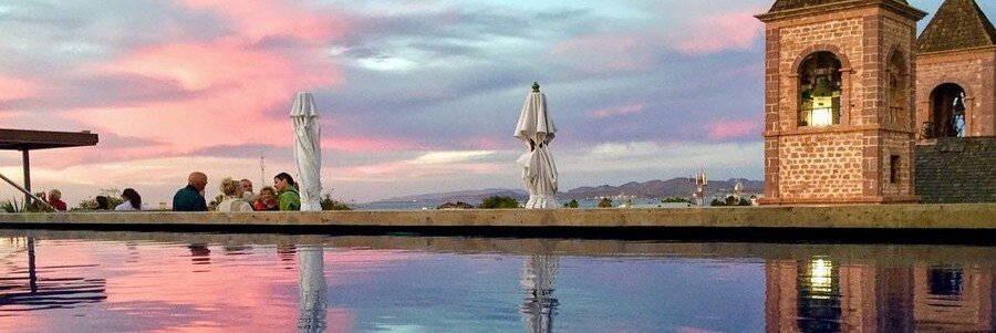 Rooftop pool at Hotel Catedral La Paz with guests relaxing at sunset, overlooking a historic stone cathedral tower and colorful sky with city and mountain views in the distance.
