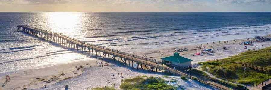 A long wooden pier extends into the ocean at sunset, with a sandy beach and people nearby.