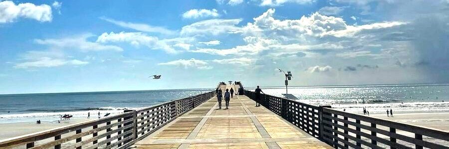 Jacksonville Beach Pier stretching into the Atlantic Ocean on a sunny day with beachgoers walking along the boardwalk.