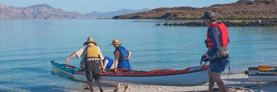 People launching a kayak into the calm, clear waters of Bahía Concepción near Mulegé, Mexico, with desert hills and a quiet beach setting