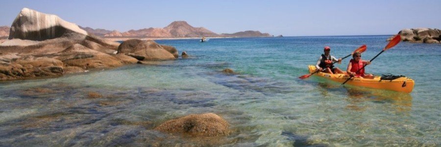 Couple kayaking through clear turquoise water near rocky coastline in San José del Cabo with scenic Baja desert mountains in the background.