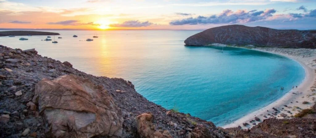 A breathtaking sunset view over Balandra Beach in La Paz, Mexico, featuring a curved white sand shoreline, turquoise bay waters, anchored boats, and surrounding desert hills under a colorful sky