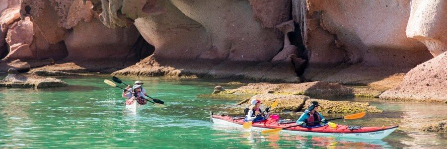 Two kayakers paddling through calm turquoise water near unique rock formations at Balandra Beach in La Paz, Mexico, surrounded by smooth sandstone cliffs and shallow coastal waters